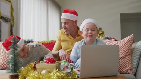Kaukasischer-Vater-Und-Zwei-Söhne-Mit-Weihnachtsmützen,-Die-Während-Der-Weihnachtszeit-Einen-Videoanruf-Auf-Dem-Laptop-Führen