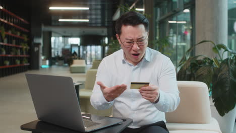 angry chinese mature businessman holding credit bank card sitting at modern workplace in office