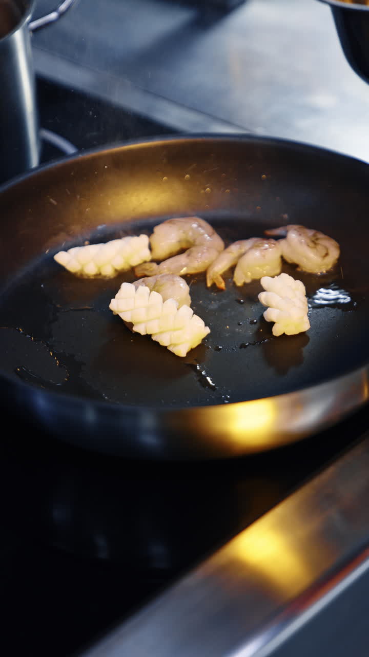 A frying-pan on the stove with some oil in it. Unrecognized cook puts some seafood to fry and moves the pieces with spatula. Close up. Vertical video.