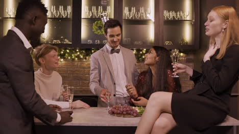 group of friends dressed in elegant clothes celebrating the new year's party, they stand around the bar counter while drinking champagne and eating grapes