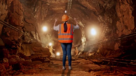 full body back view of a female engineer with safety helmet raising her hands celebrating while working in underground mine tunnel