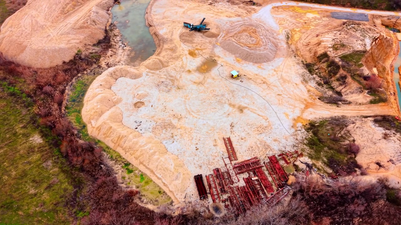 Drone view of quarry lake shore with stacked steel frames. Oblique perspective of red steel structure piles on sandy peninsula, teal lake water, green strip and bare deciduous trees.