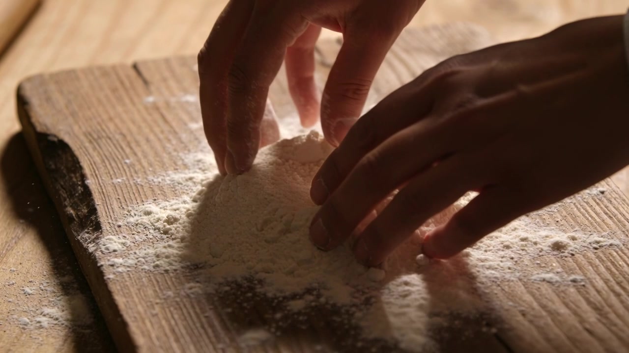 Pressing hands shaping flour mound on wood board, white cuff pinching ingredient for dough mixing