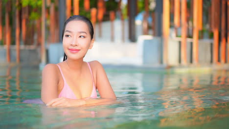 수영장에 있는 매력적인 젊은 몸매의 여성이 카메라 밖의 무언가를 보기 위해 몸을 돌릴 때 계단 가장자리를 따라 몸을 기울입니다.