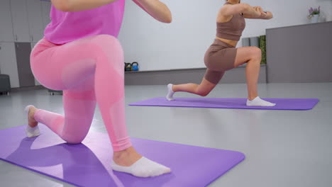 partial view of two women performing lunges on yoga mats in a well-decorated gym, they are engaged in a synchronized workout routine, focusing on flexibility and strength training