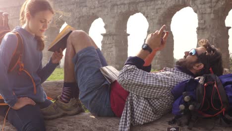 happy young couple backpackers tourists sitting lying on a log trunk reading book using smartphone in front of ancient roman aqueduct ruins in parco degli acquedotti park in rome at sunrise romantic with sleeping bag slow motion