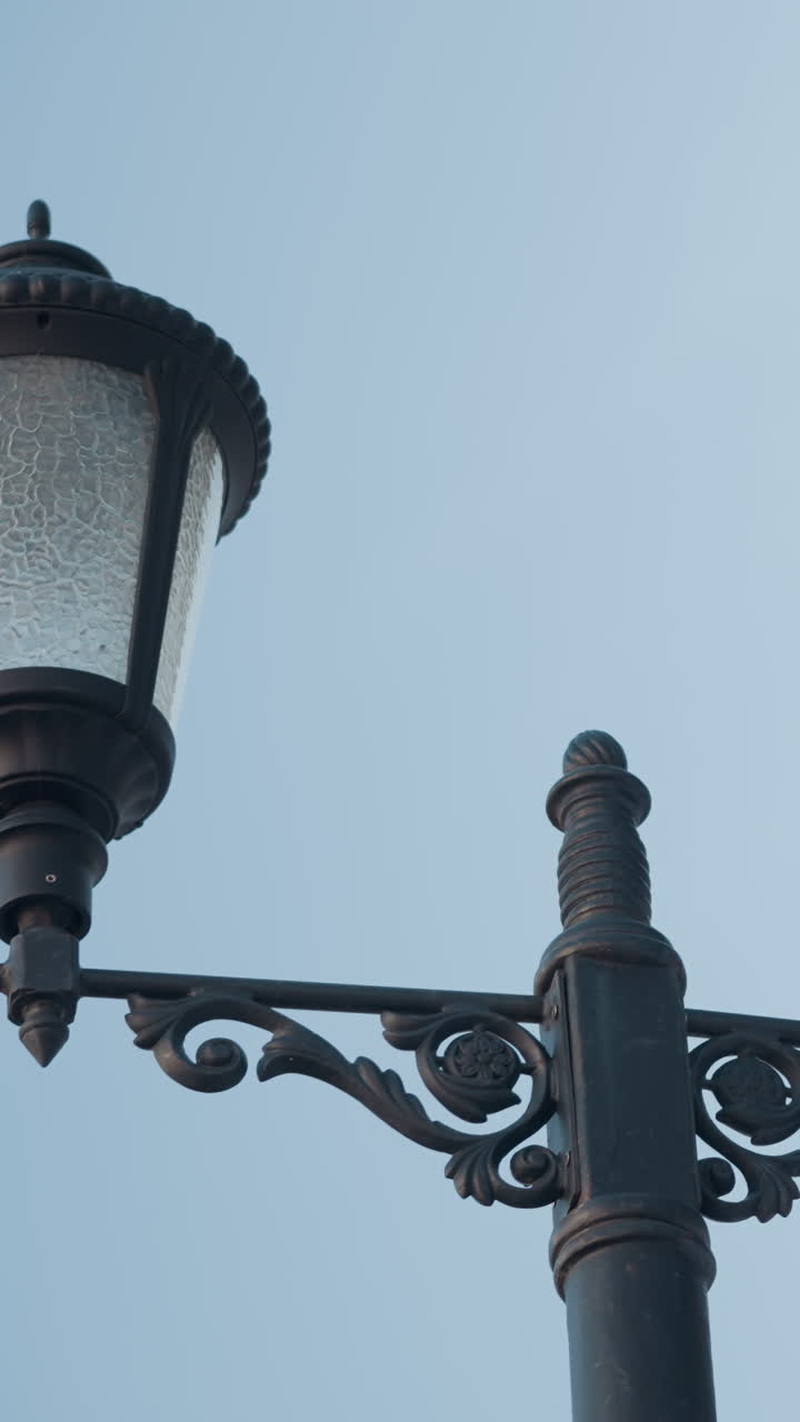Close-up of an ornate black double-lamp post with intricate designs, highlighting textured glass shades against a clear blue sky and partial view of a beige patio canopy