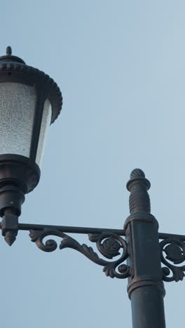 primer plano de un ornamentado poste de doble lámpara negra con diseños intrincados, destacando sombras de vidrio texturizadas contra un cielo azul claro y una vista parcial de un dosel de patio beige
