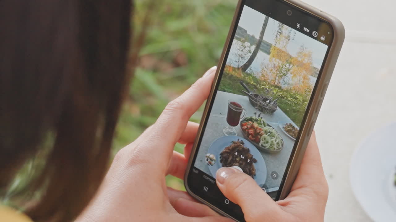Closeup of meal on mobile device during camping. Touchscreen captures dinner setting with salad and mug. Photograph showing hands holding phone to capture outdoor dining scene with golden light
