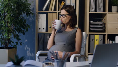 pregnant businesswoman in glasses caressing her belly and drinking tea sitting on a chair in the office