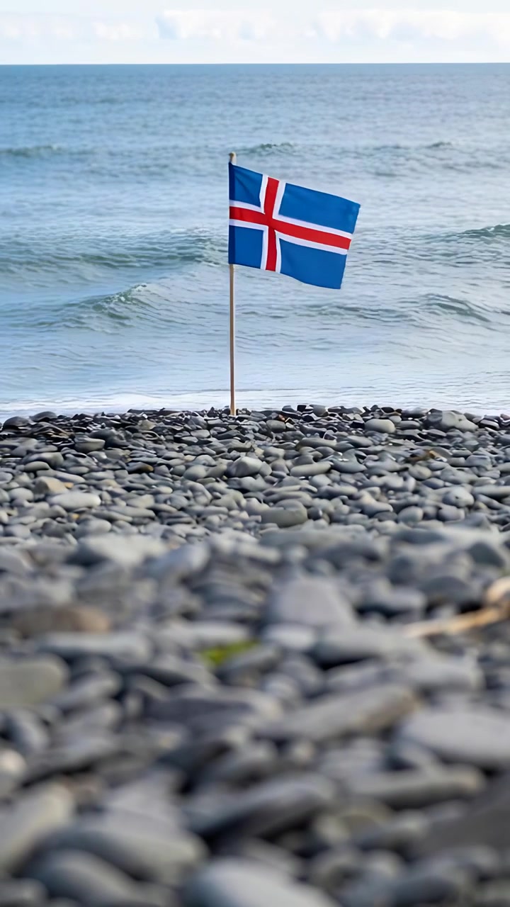 Vertikal video: Bris rører islandsk flagg på tynn stang som flagrer ved småstein, bølger ruller inn