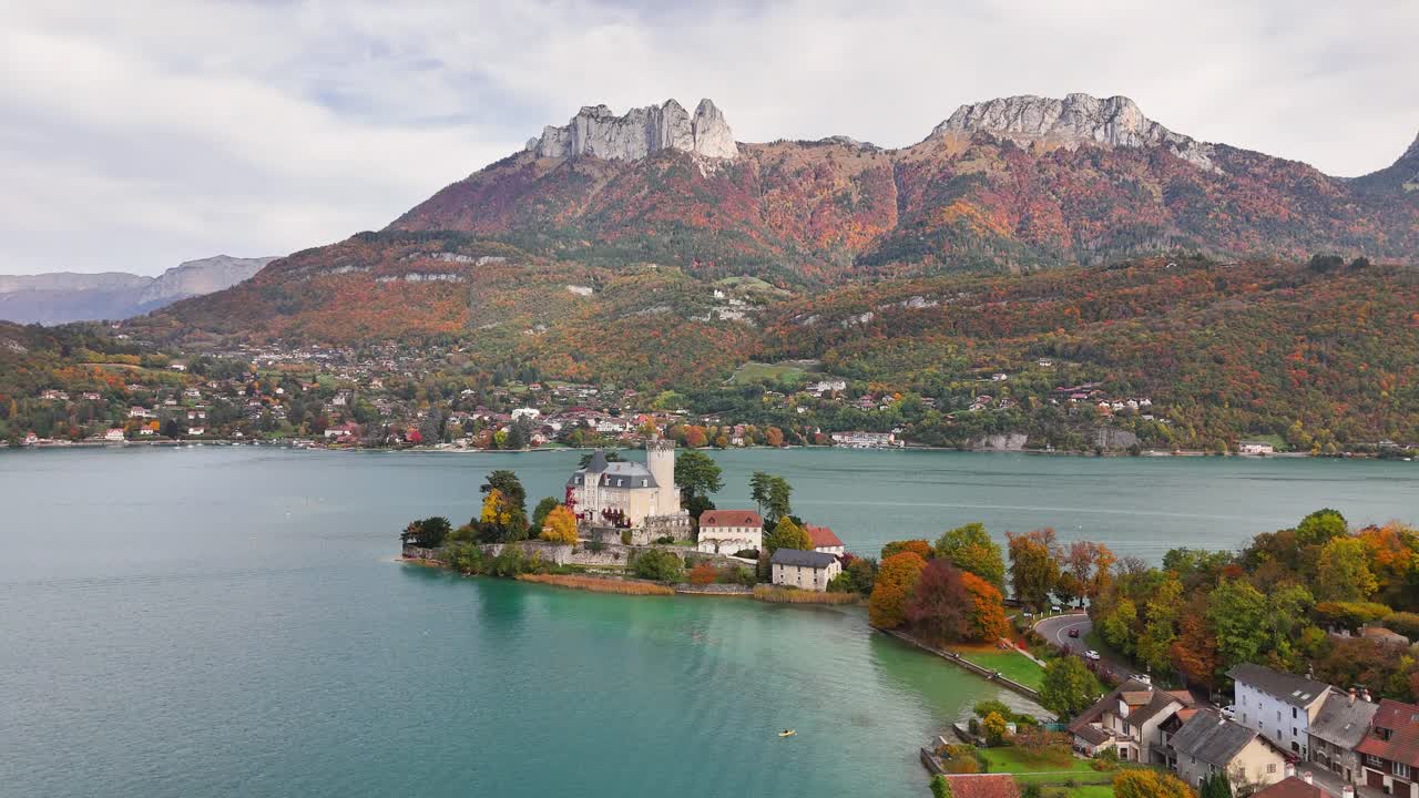 Chateau de Duingt in France stands on a narrow peninsula surrounded by turquoise lake waters, vibrant autumn foliage, and mountain peaks, with nearby village houses nestled along the serene shoreline