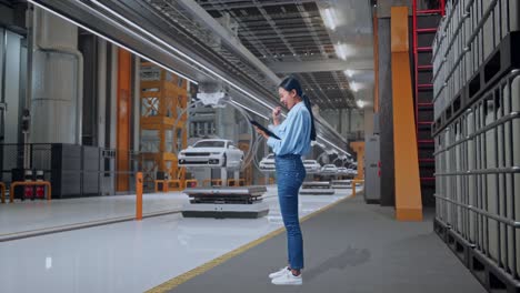 vista lateral completa del cuerpo de una trabajadora profesional asiática de pie con su tableta en una fábrica de automóviles ev, instalación industrial de alta tecnología, levanta el puño con el objetivo de gritar
