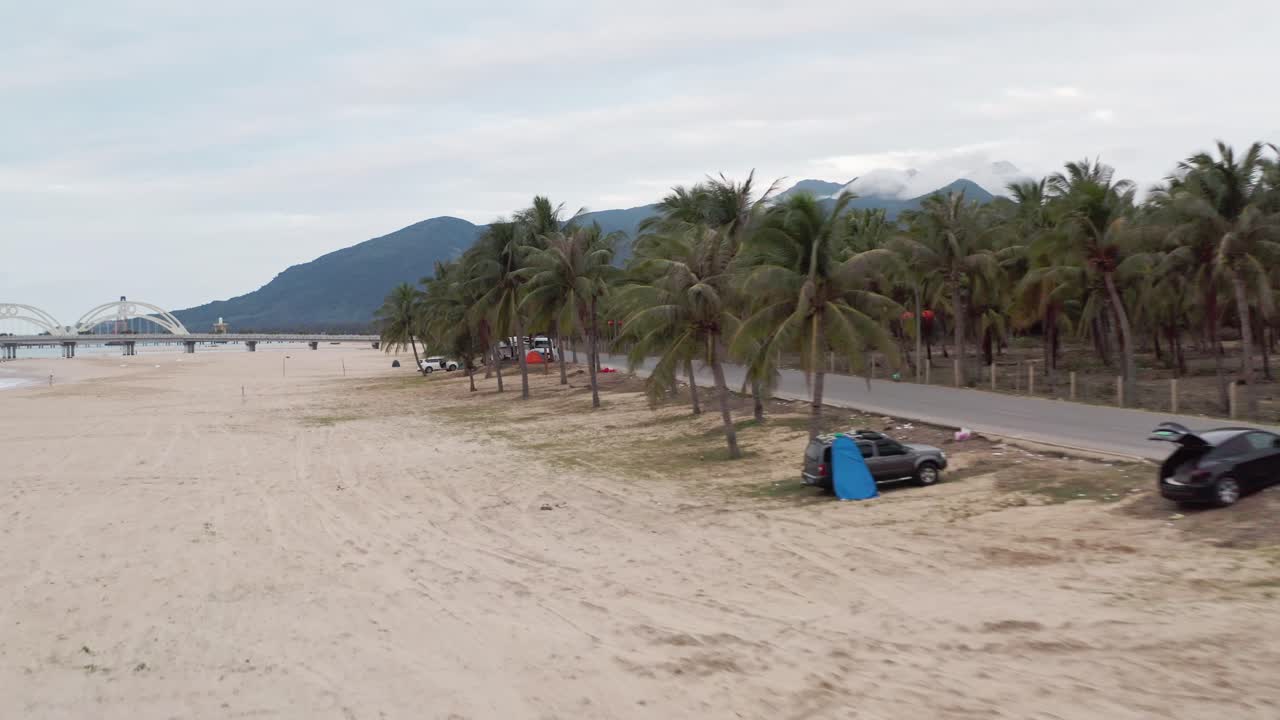 autocaravanas y vehículos recreativos estacionados bajo palmeras en la carretera lateral de la playa tropical, con montañas cubiertas de nubes en el fondo