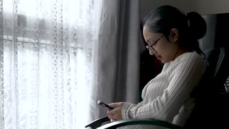 Young-British-Asian-Woman-in-sweater-using-smartphone-by-window,-natural-light,-serene-mood,-swiveling-on-chair