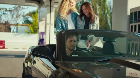 guy and two girls arrived in a gray convertible to a gas station. the guy gets out of the driver's seat and fills up the car