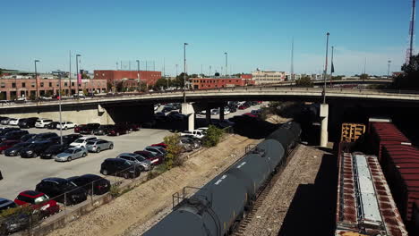 El-Video-De-Un-Dron-Muestra-Un-Tren-De-Carga-Que-Transporta-Mercancías-Por-Tierra-A-Través-De-Vías,-Así-Como-Muchos-Autos-Estacionados-Al-Costado-Y-Un-Paso-Elevado