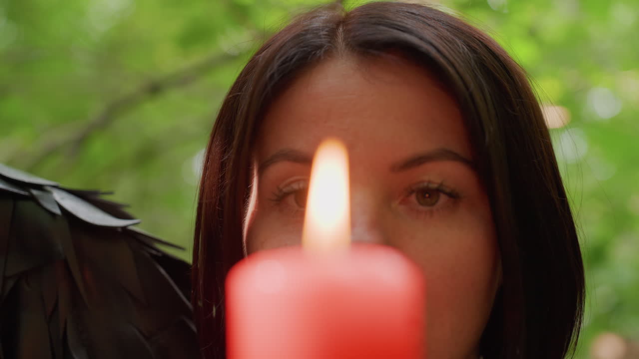 Koyu renk saçlı bir kadın tarafından tutulan kırmızı mum alevinin aşırı yakın çekimi, yumuşak parıltıya ve bulanık yeşillik arka plana odaklanarak, meditatif sakin bir mistik atmosfer yaratıyor