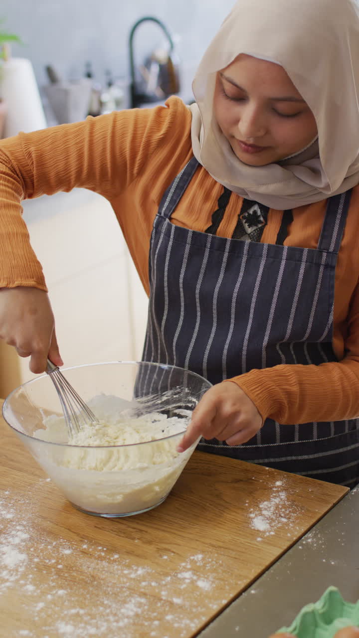 Video vertikal seorang wanita birasial berhijab yang bahagia sedang memanggang di dapur, mengaduk adonan kue di mangkuk.