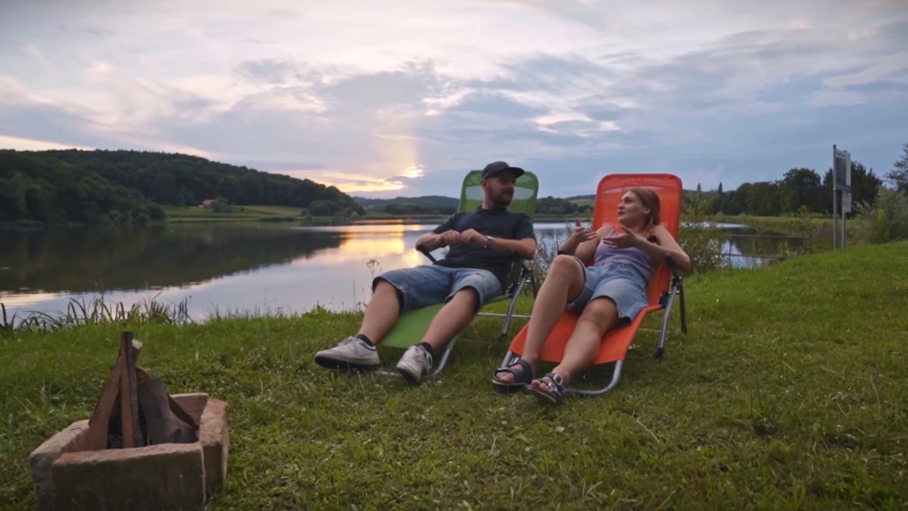 Coppia seduta su sedie da esterno vicino a un lago calmo al tramonto, che parla e si gode la tranquilla atmosfera del campeggio e i suggestivi dintorni naturali a Savci, Slovenia
