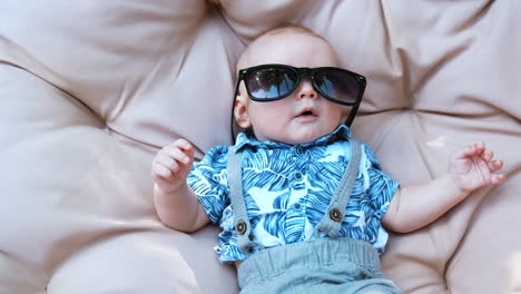 funny baby boy wearing mom's sunglasses. kid wearing a blue shirt and shorts lies on a soft pillow outdoors.
