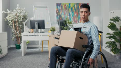 disabled ambitious man in shirt with asian korean beauty sits in wheelchair at desk in the company on knees holding cardboard box with packed things, gets fired from job unable to fulfill his duties