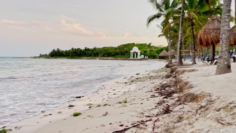 Sliding-left-shot-of-a-beautiful-beach-in-Tulum-near-Cancun-Mexico-with-small-waves,-palm-trees,-blue-sky-and-some-clouds