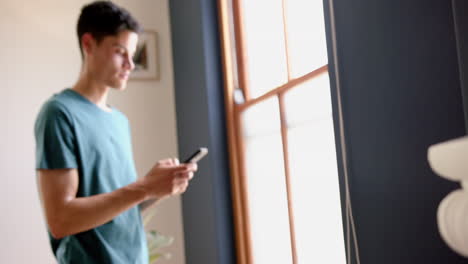 focused biracial man standing by sunny window using smartphone at home, copy space, slow motion