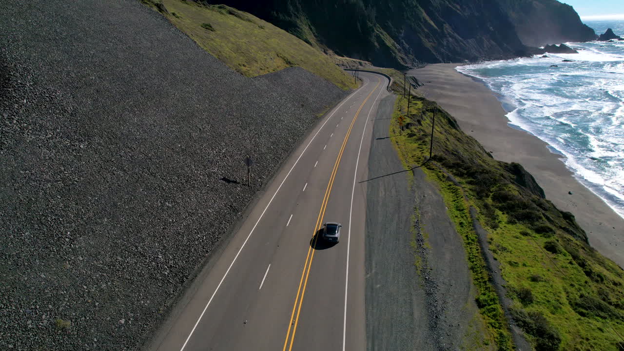 A car driving along the Oregon Coast Highway with mountains and the ocean in the background.