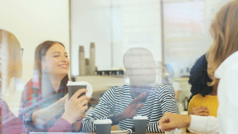 camera focuses on a multiethnic group of friends through the window talking while drinking coffe sitting at a table in a cafe