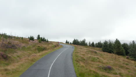 Fly-over-vintage-convertible-car-on-road-rising-up-to-hill.-Revealing-cyclist-slowly-climbing.-Autumn-weather-with-fog-in-valley.-Ireland