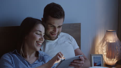 young caucasian couple sitting the bed and watching a video on the tablet device at night