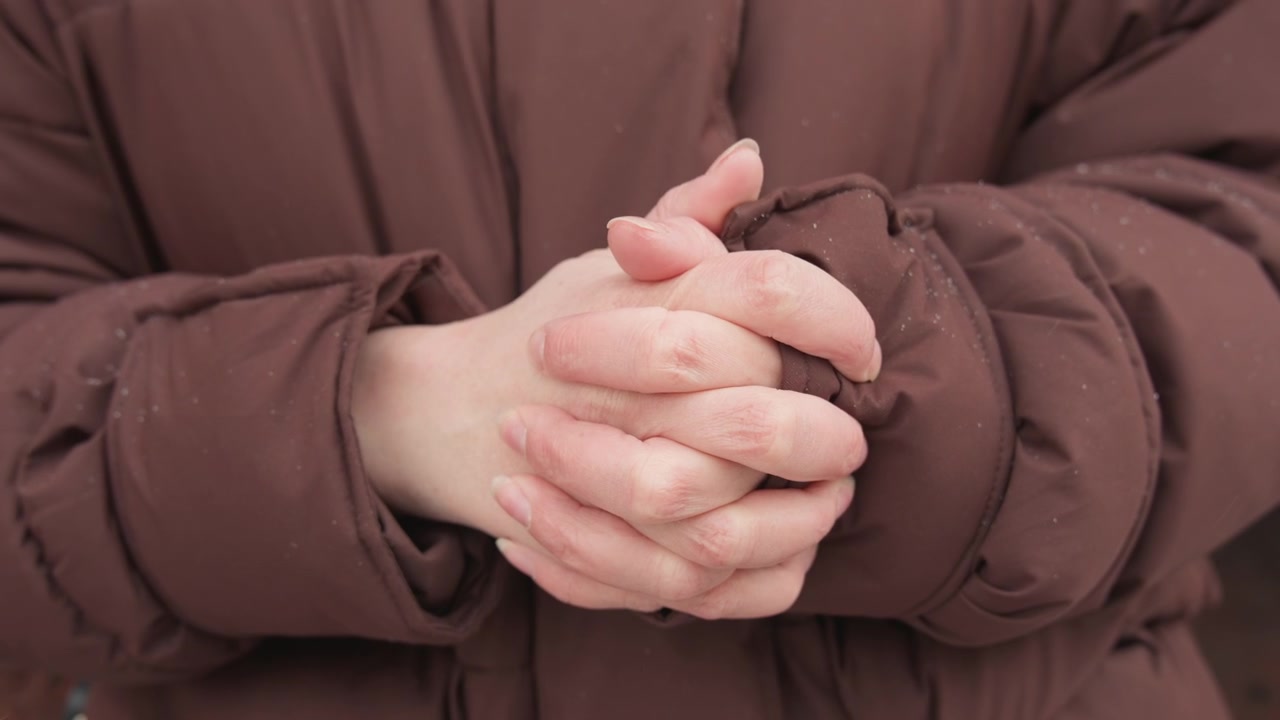 Tampilan dekat seseorang mengenakan jaket puffer berlapis cokelat dengan tangan bersedekap dan tangan terselip di lengan baju, menunjukkan postur mencari kehangatan yang protektif saat bintik-bintik putih kecil menempel pada kain bertekstur