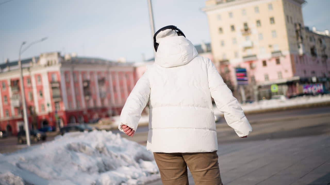 Back view of young woman twirling in white winter jacket and boots on snowy urban plaza, arms extended embracing winter breeze while passing cars and buildings frame lively outdoor dance moment
