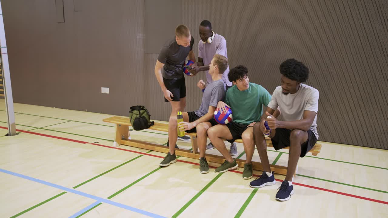 Fünf Männer kommen von rechts, setzen sich in der Turnhalle zwischen den Spielzügen auf eine Bank und reichen sich Volleybälle und Flaschen zu.