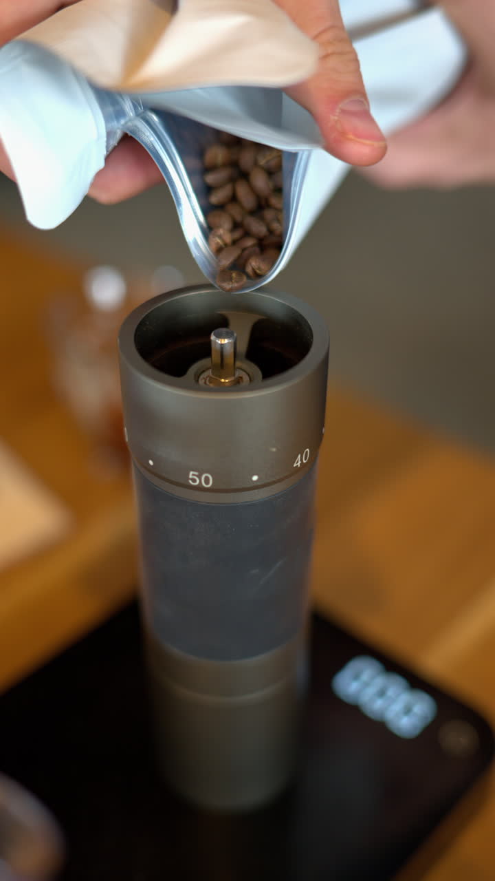 Close up of a man pouring roasted coffee beans from a white bag into a manual coffee grinder. Vertical