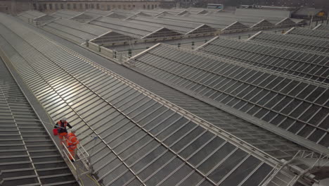 Pan-up-from-two-workmen-in-orange-high-visibility-vests-cleaning-the-roof-of-Waverley-railway-station-to-Edinburgh-city-on-a-sunny-day,-Scotland