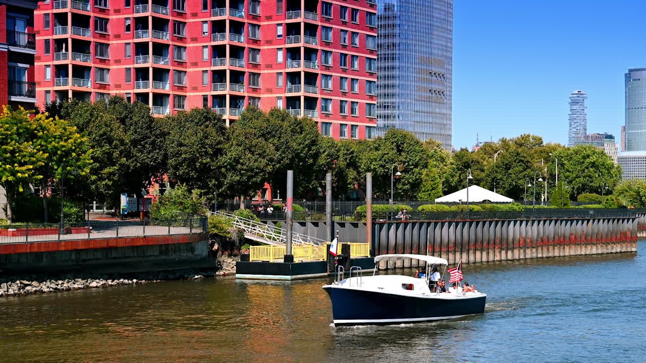Jersey City, USA, 1 July 2025: Motorboat with group of people approaches the piers. View on the green quay of Jersey City. Skyscrapers of Manhattan at backdrop
