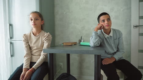 two bored schoolchildren sitting at table in modern kitchen, looking tired and uninterested, surrounded by books and school supplies, possibly doing homework or waiting for meal or parents