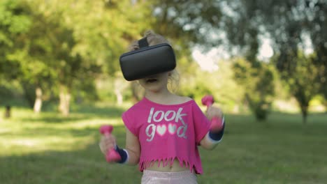 athletic child girl in vr headset helmet making fitness workout exercises with dumbbells in park