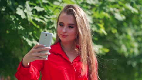 attractive model with a mobile phone outdoors. beautiful woman doing selfie in the street on green tree background. lovely girl is recording video of herself on a phone.