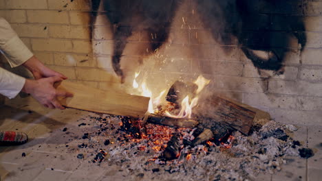 Male's hands split the heat with a log in the fire indoors. Close-up hands of a man putting wood into fire in fireplace and then warming up his cold hands. Christmas time.