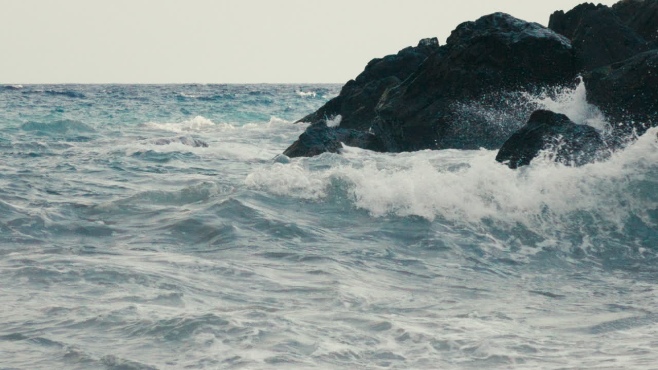 Wide slow motion shot of a rising ocean wave approaching rugged coastal rocks. The choppy water captures raw energy, force and natural rhythm