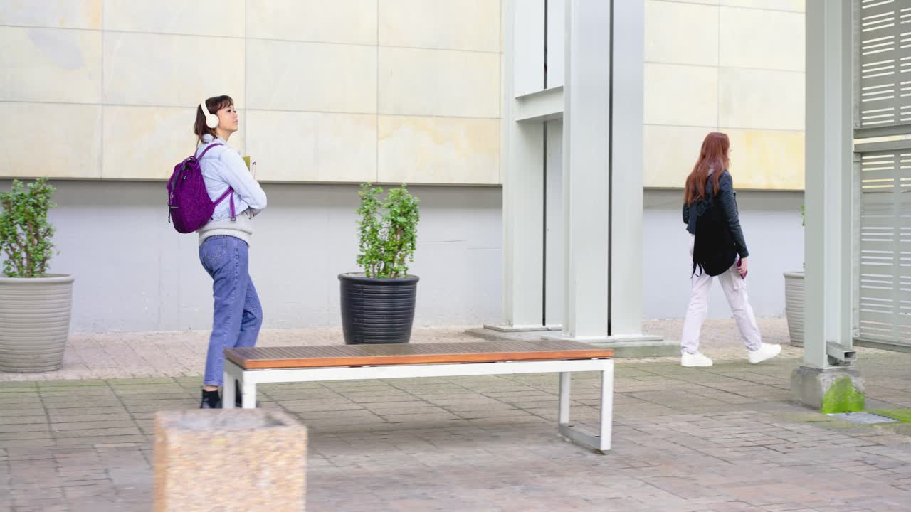 Due donne che attraversano una piazza, la rossa in testa, seguita dalla più giovane che entra a sinistra controllando il telefono.