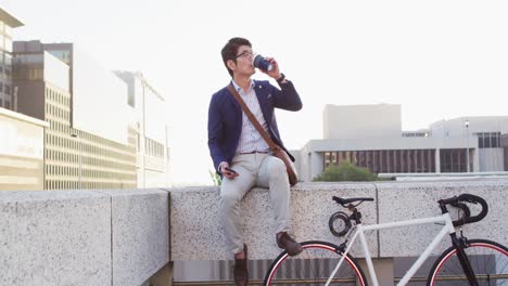 Asian-man-drinking-coffee-and-using-smartphone-while-sitting-on-the-roof-of-corporate-park