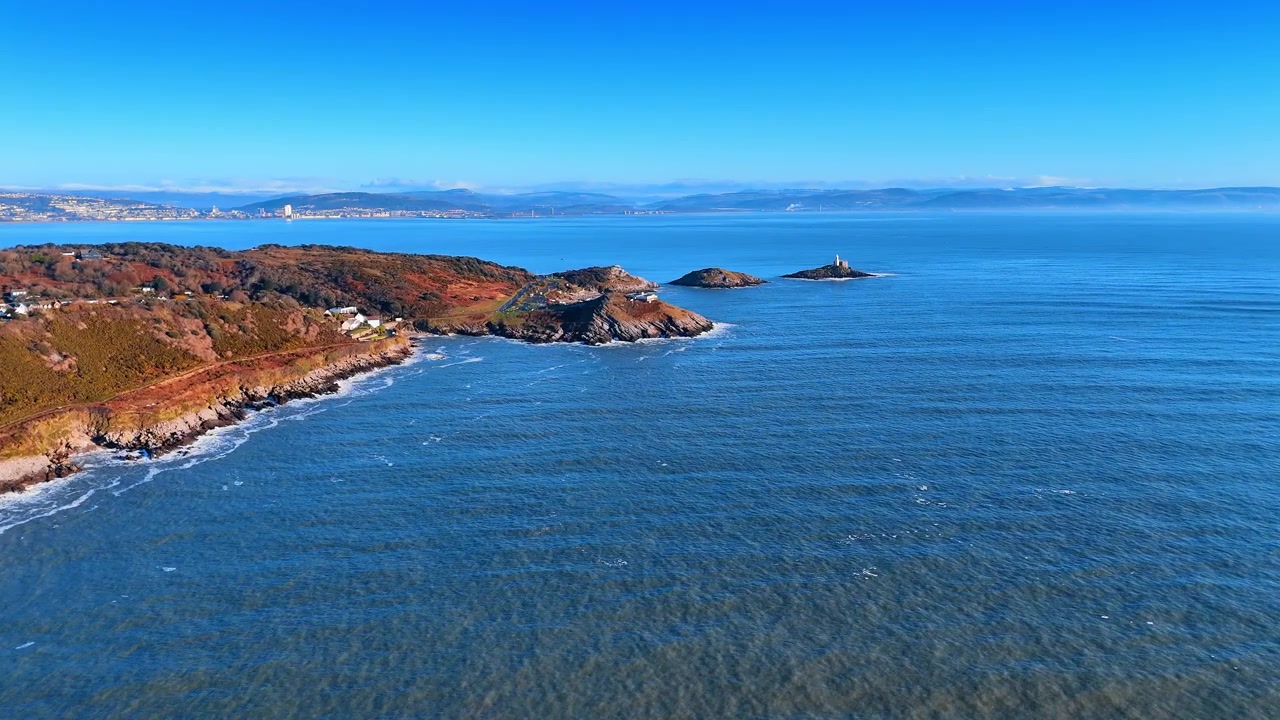 Veduta aerea di un promontorio gallese con spiaggia e villaggio costiero sullo sfondo. Un promontorio verde incontra il mare accanto a una cala sabbiosa, mentre un villaggio costiero e capanne sulla spiaggia si estendono lungo la costa nell'entroterra.