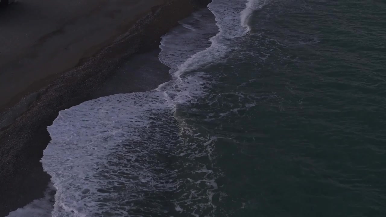 High angle aerial shot of green sea waves gently washing onto a dark sand and pebble beach shore creating white foam patterns under soft twilight lighting, ideal for nature background concept.
