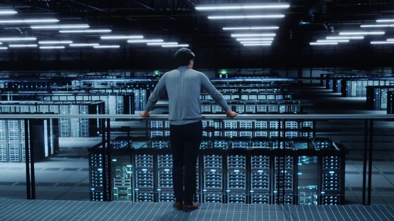 Datacenter-ingeniør kigger ned fra en balkon på en serverfarm cloud computing-specialistfacilitet. Multietnisk asiatisk systemadministrator arbejder med databeskyttelsesnetværk for cybersikkerhed.