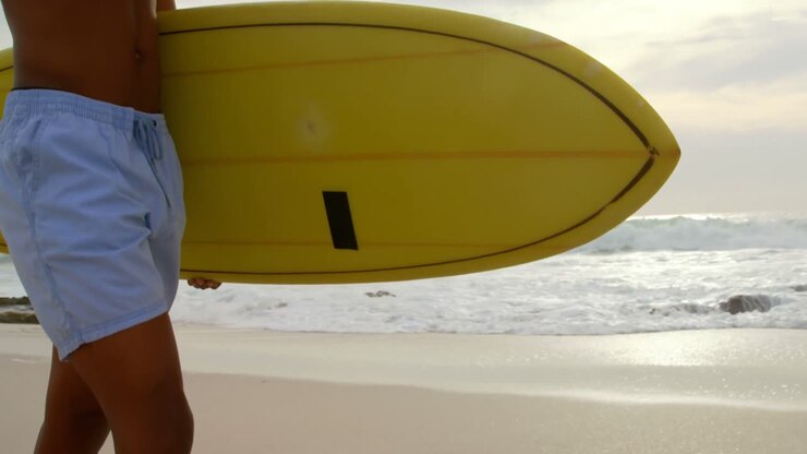 Vista laterale di surfista afroamericano che cammina con la tavola da surf sulla spiaggia 4k