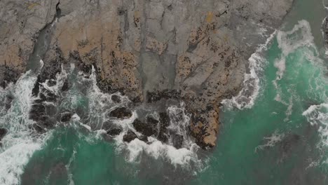 An-aerial-shot-of-the-crashing-waves-at-Pentire-Point-near-Polzeath-beach,-the-water-is-turquoise-and-bright-during-the-summer-months